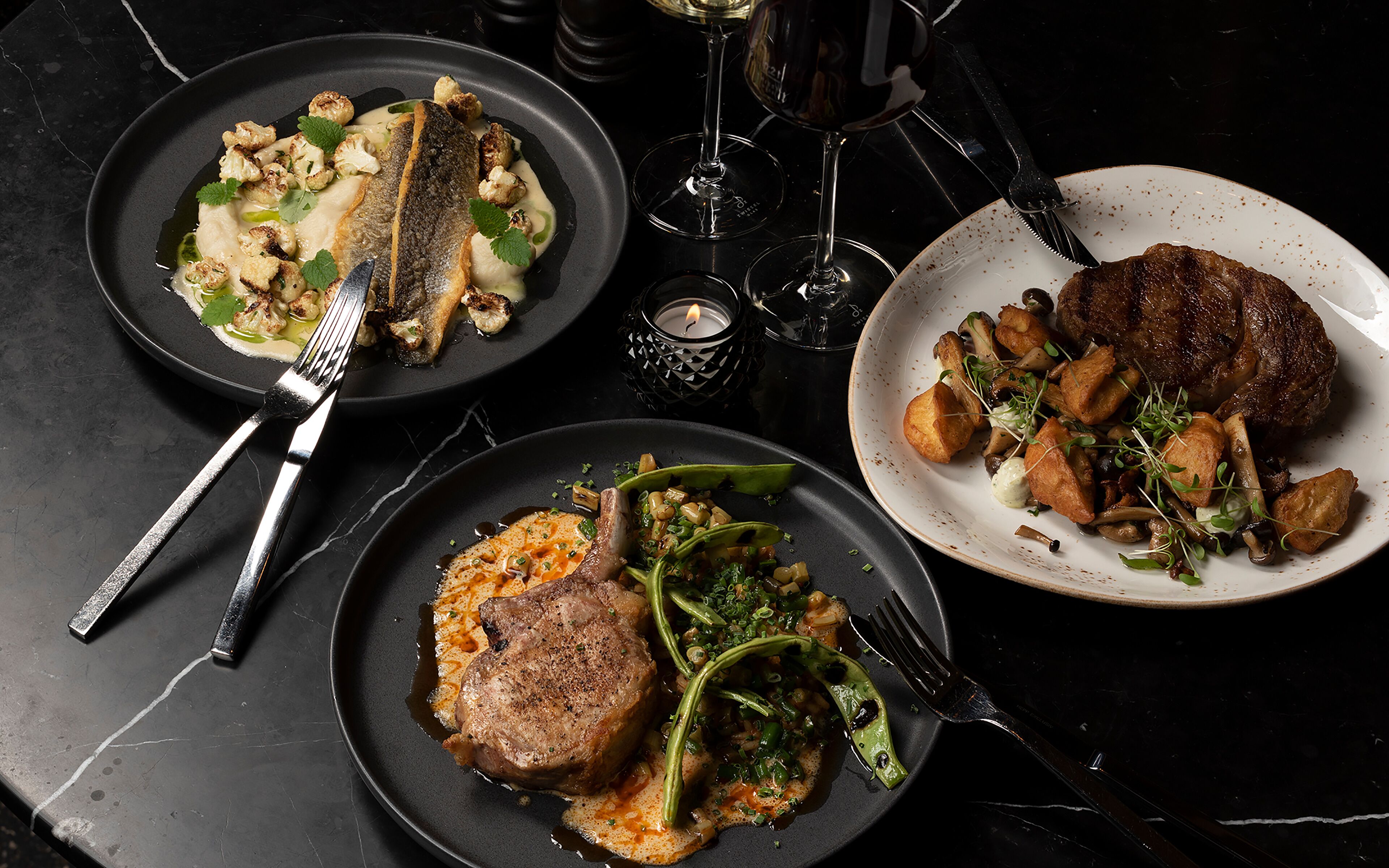 Three plates of food on a dark surface with forks and a candle.