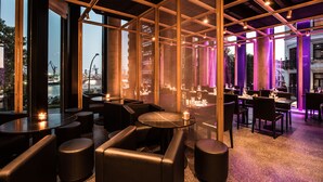 A modern interior with a bar area, seating, and large windows offering a view of the cityscape. - Empire Riverside Hotel (Hamburg)