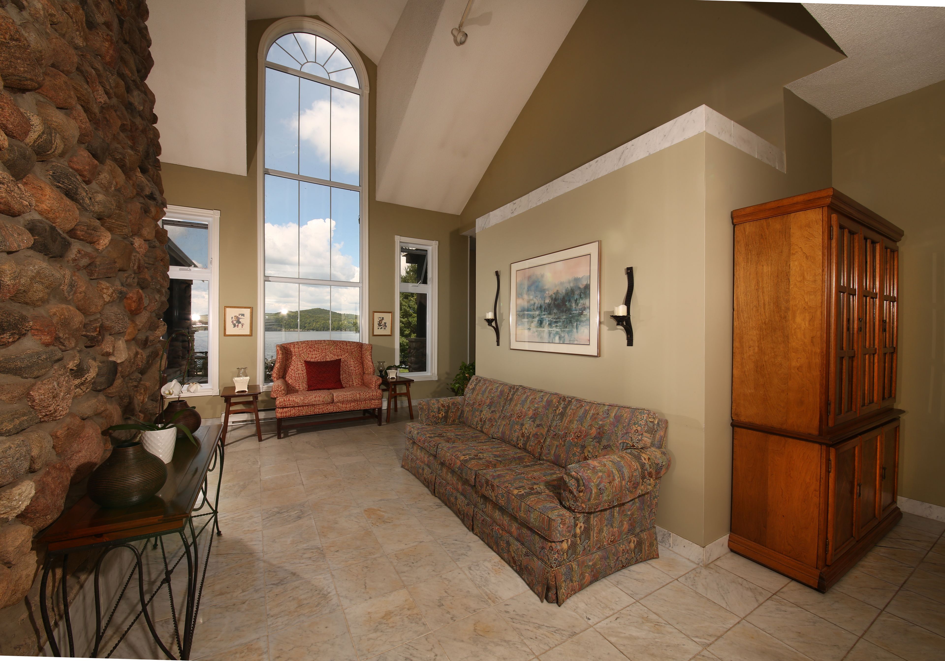 A spacious living room with a stone wall, a large window, a patterned sofa, and a wooden cabinet.