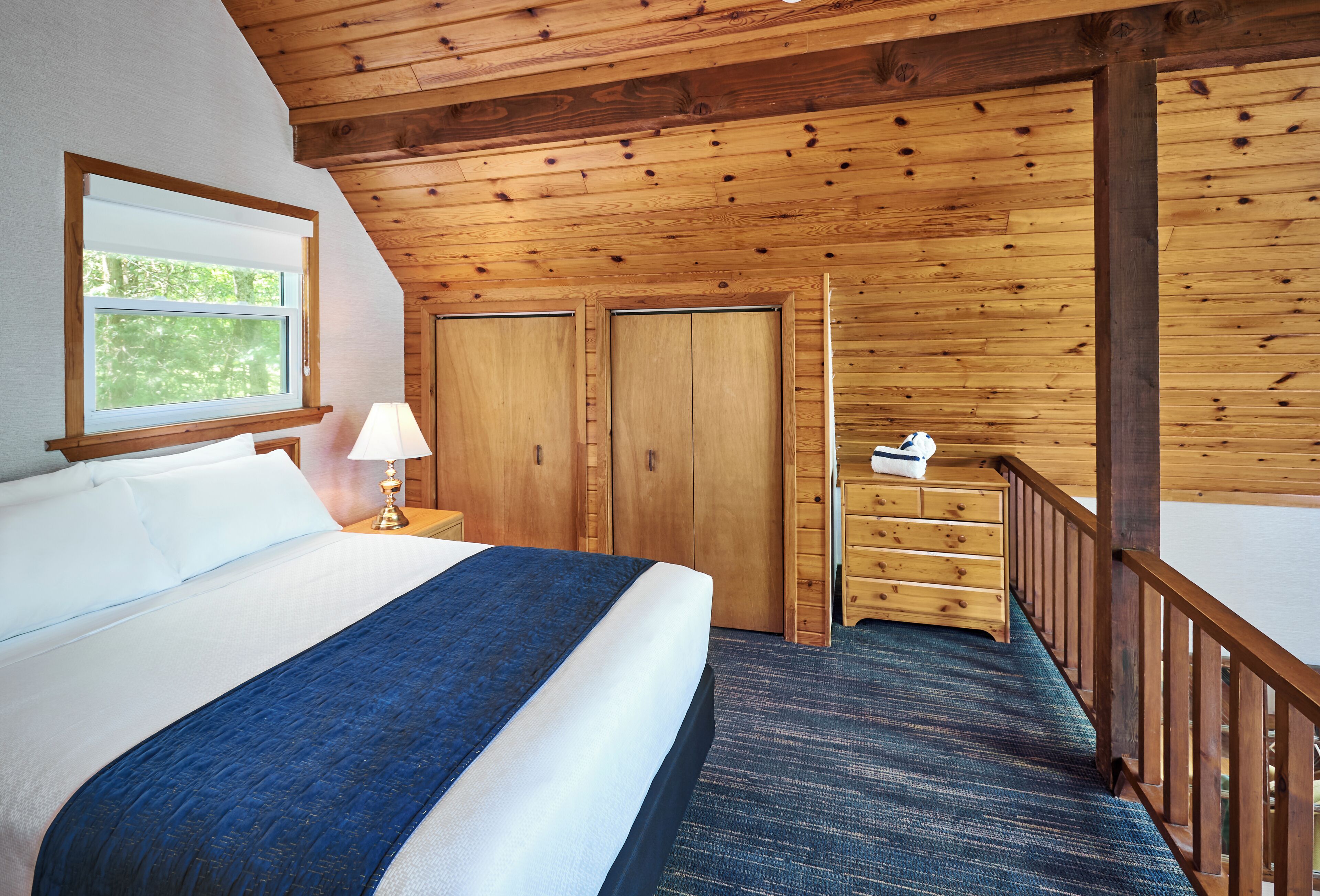 A room with a large bed, wooden ceiling, a window, a desk, and a dresser.