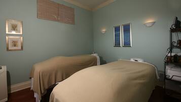 A room with two beds covered in beige linens, a wall-mounted heater, a shelf with towels, and two framed pictures on the wall.