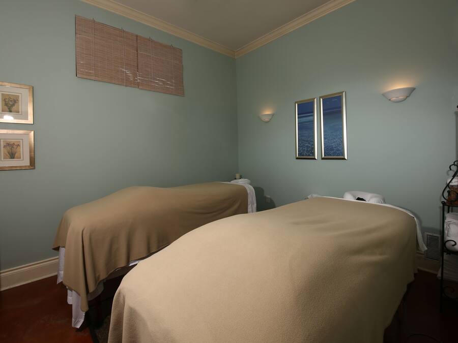 A room with two beds covered in beige linens, a wall-mounted heater, a shelf with towels, and two framed pictures on the wall.