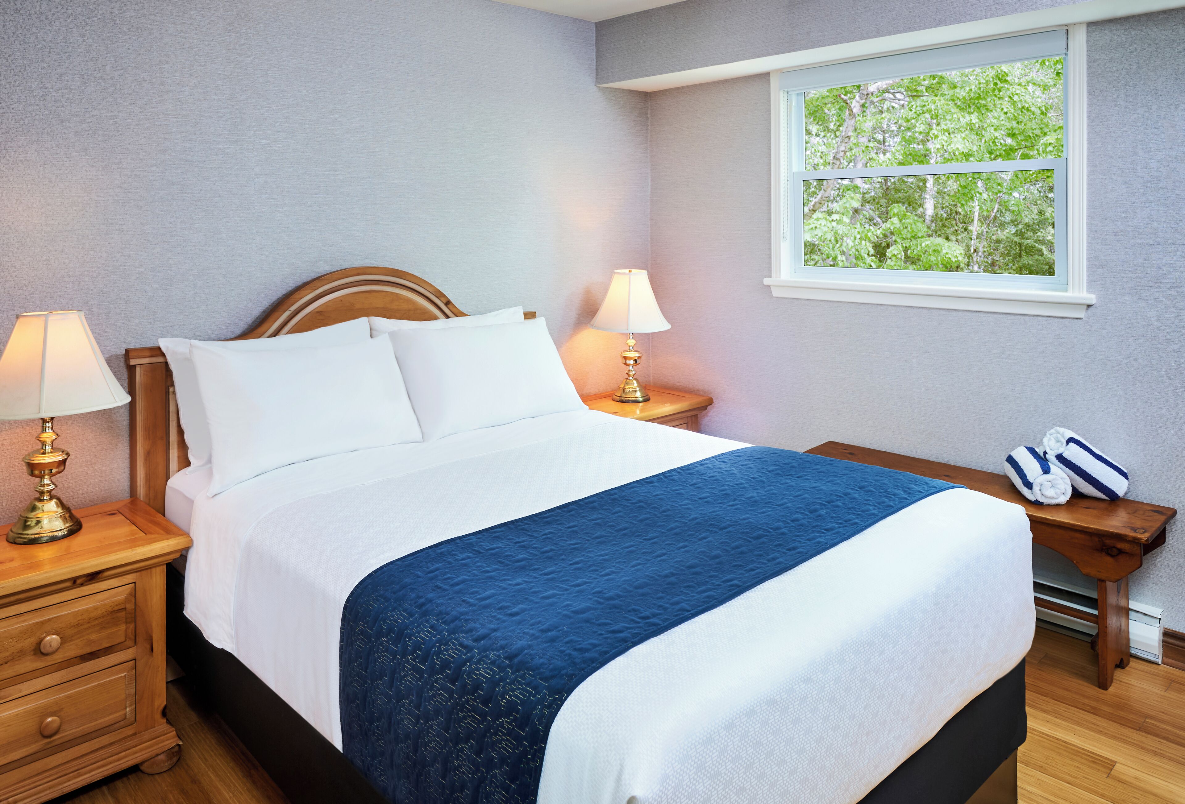A neatly made bed with a blue and white bedspread, two bedside lamps, and a window with a view of trees.