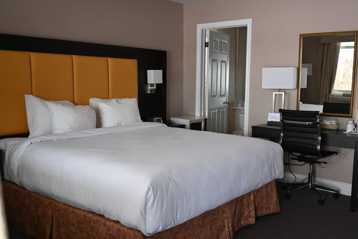 Quarto de hotel com uma cama grande, uma escrivaninha com uma cadeira, uma luminária de mesa, um espelho e uma porta que dá para outro cômodo.