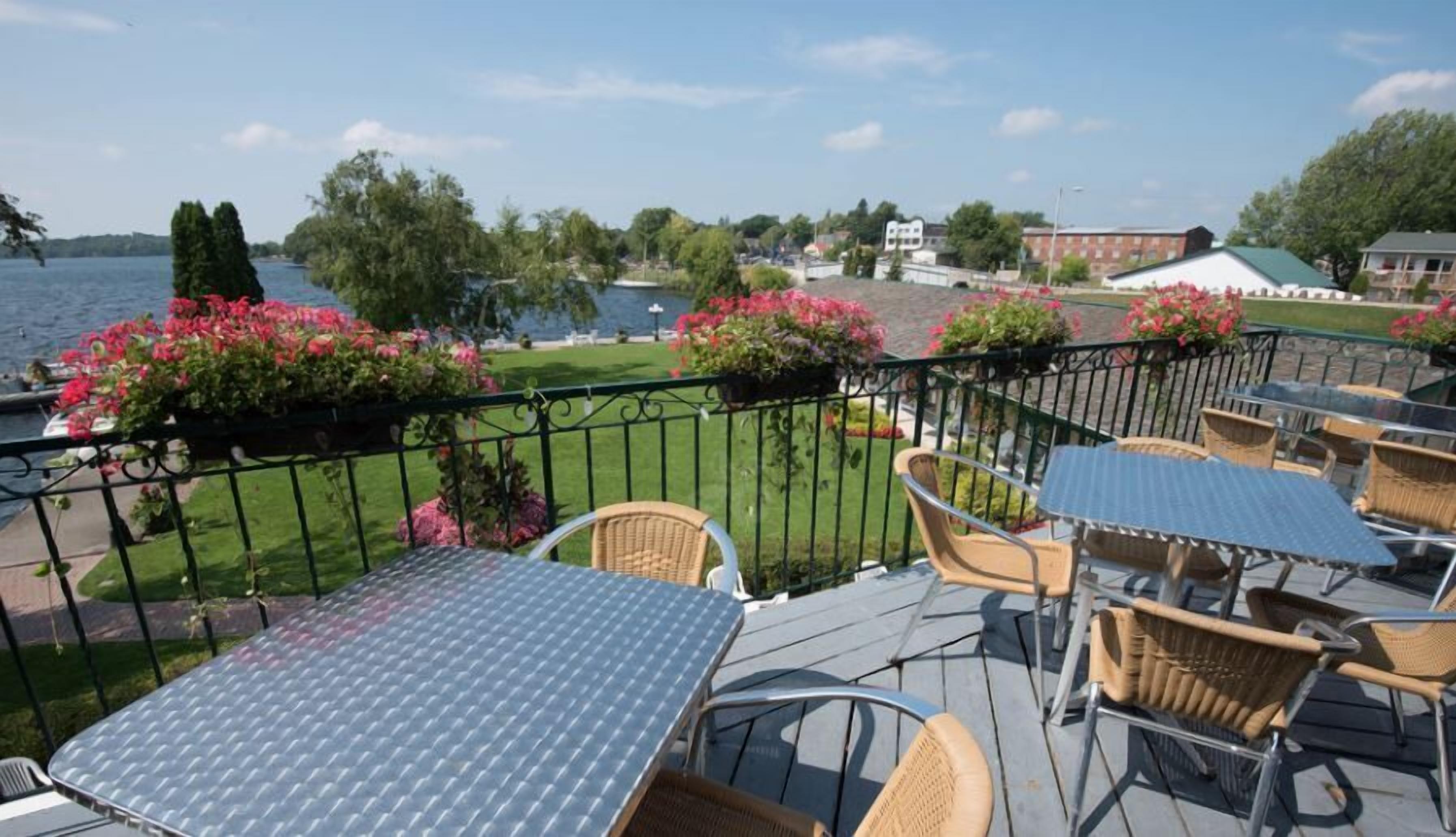 An outdoor patio with tables and chairs, overlooking a body of water and a grassy area with flowers.