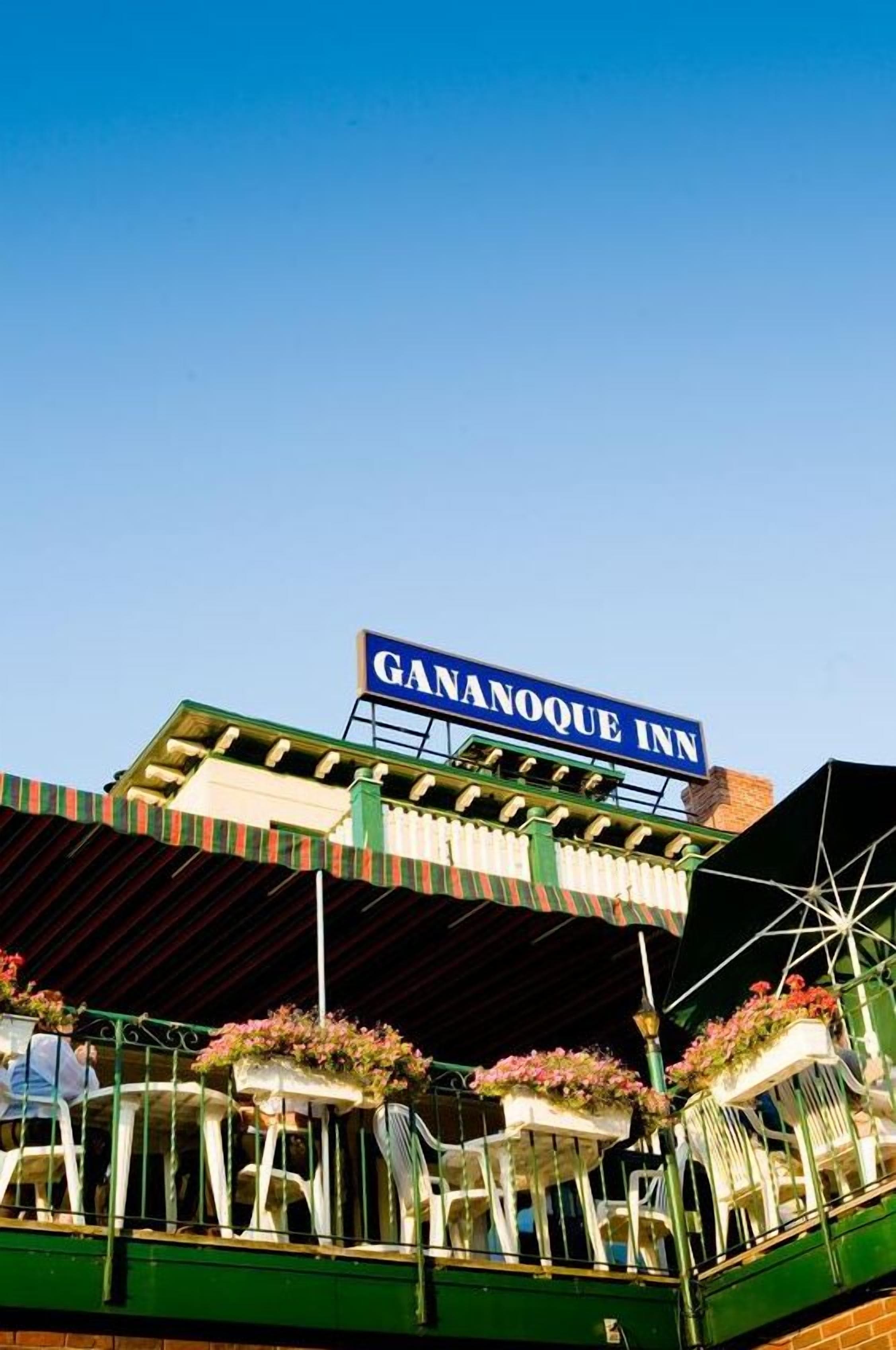A hotel named Gananoque Inn with a prominent sign, outdoor seating area with chairs and planters, and a clear blue sky in the background.