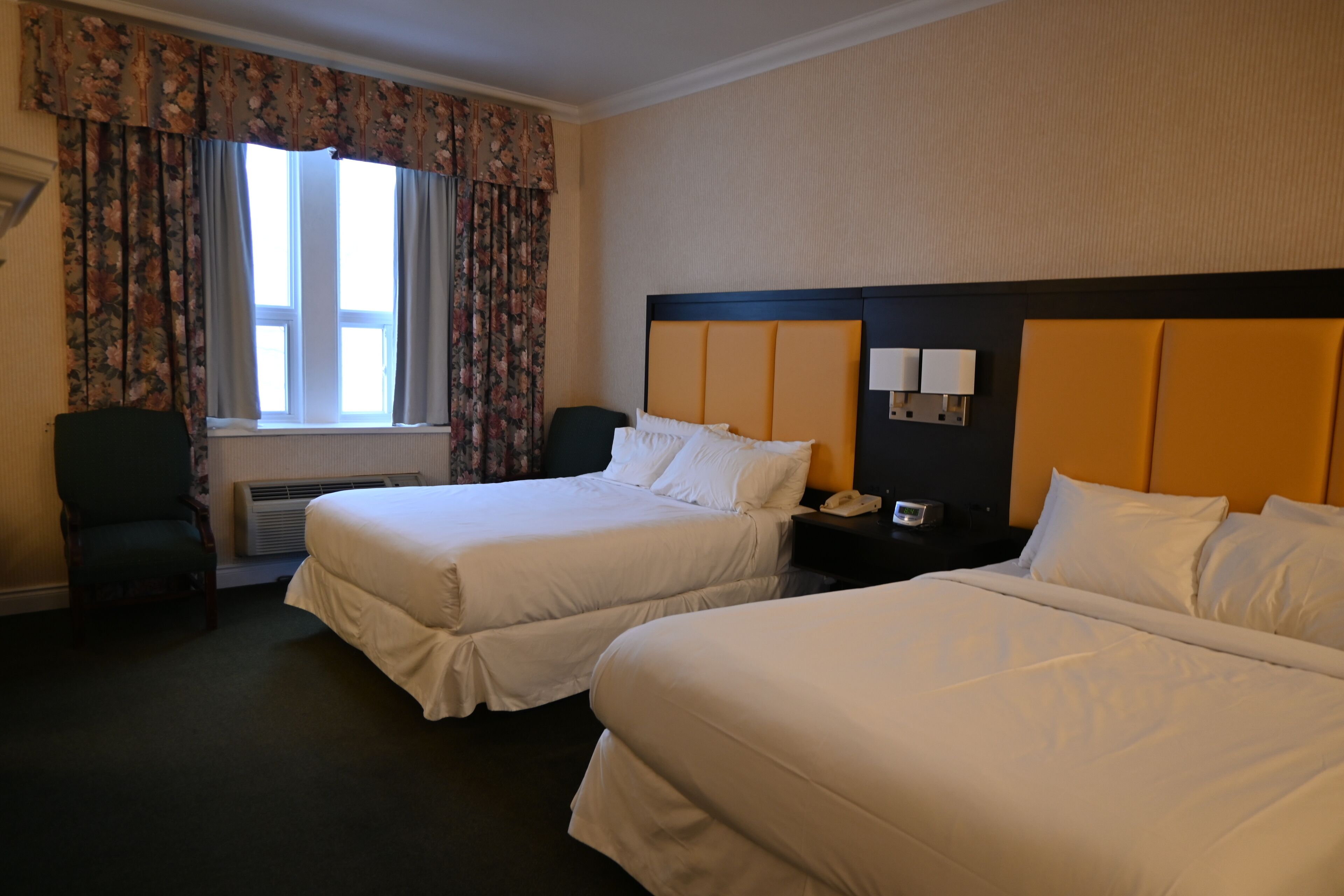 A hotel room with two beds, a chair, a nightstand with a lamp, and a window with curtains.