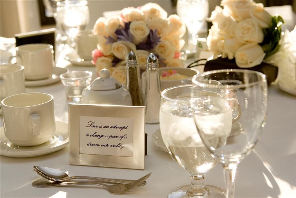 A table set with white cups, saucers, a teapot, sugar and salt shakers, a glass of water, and a card with a quote.