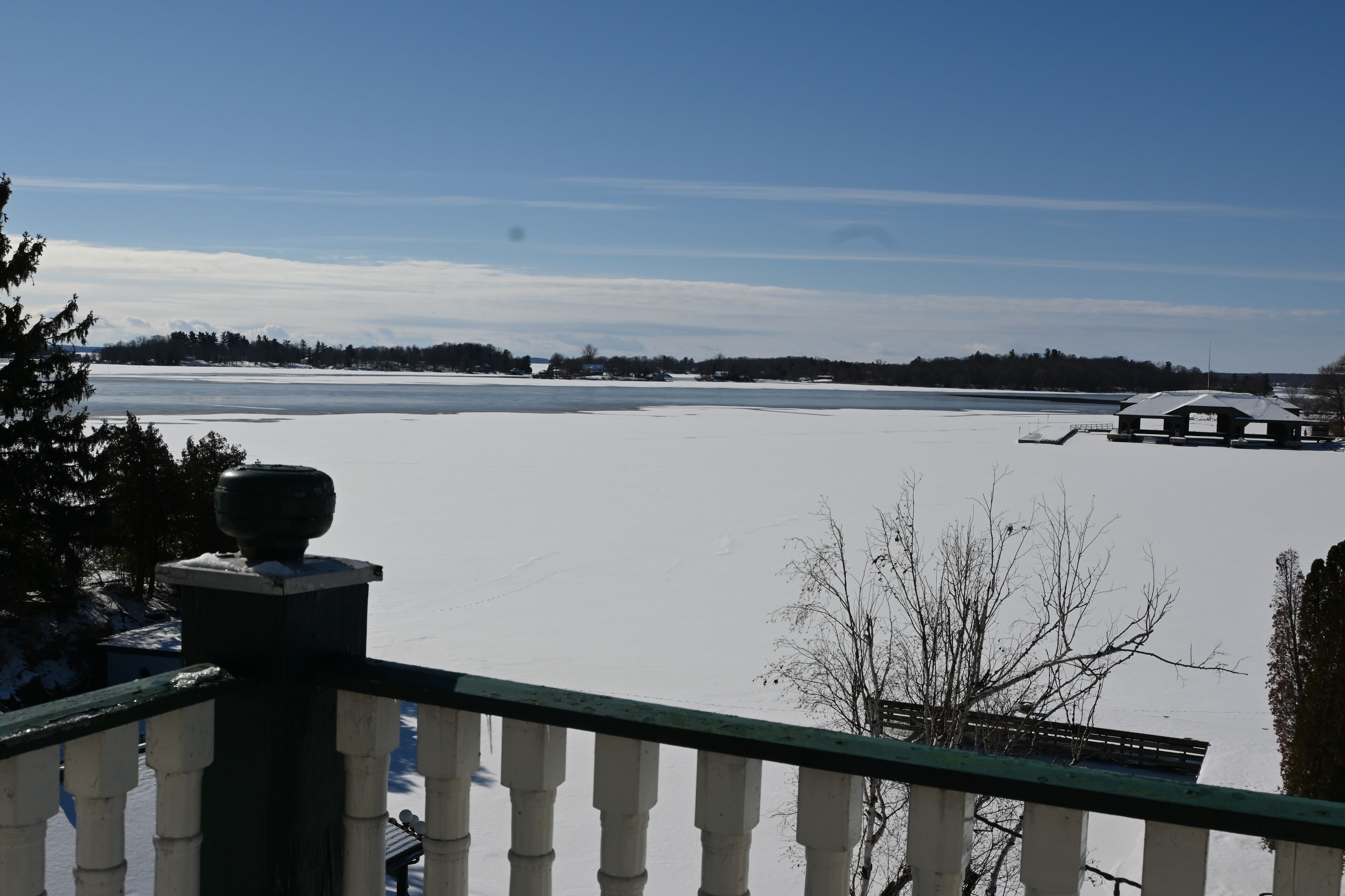 A snowy landscape with a clear view of a large body of water, a distant tree line, and a building with a green roof.