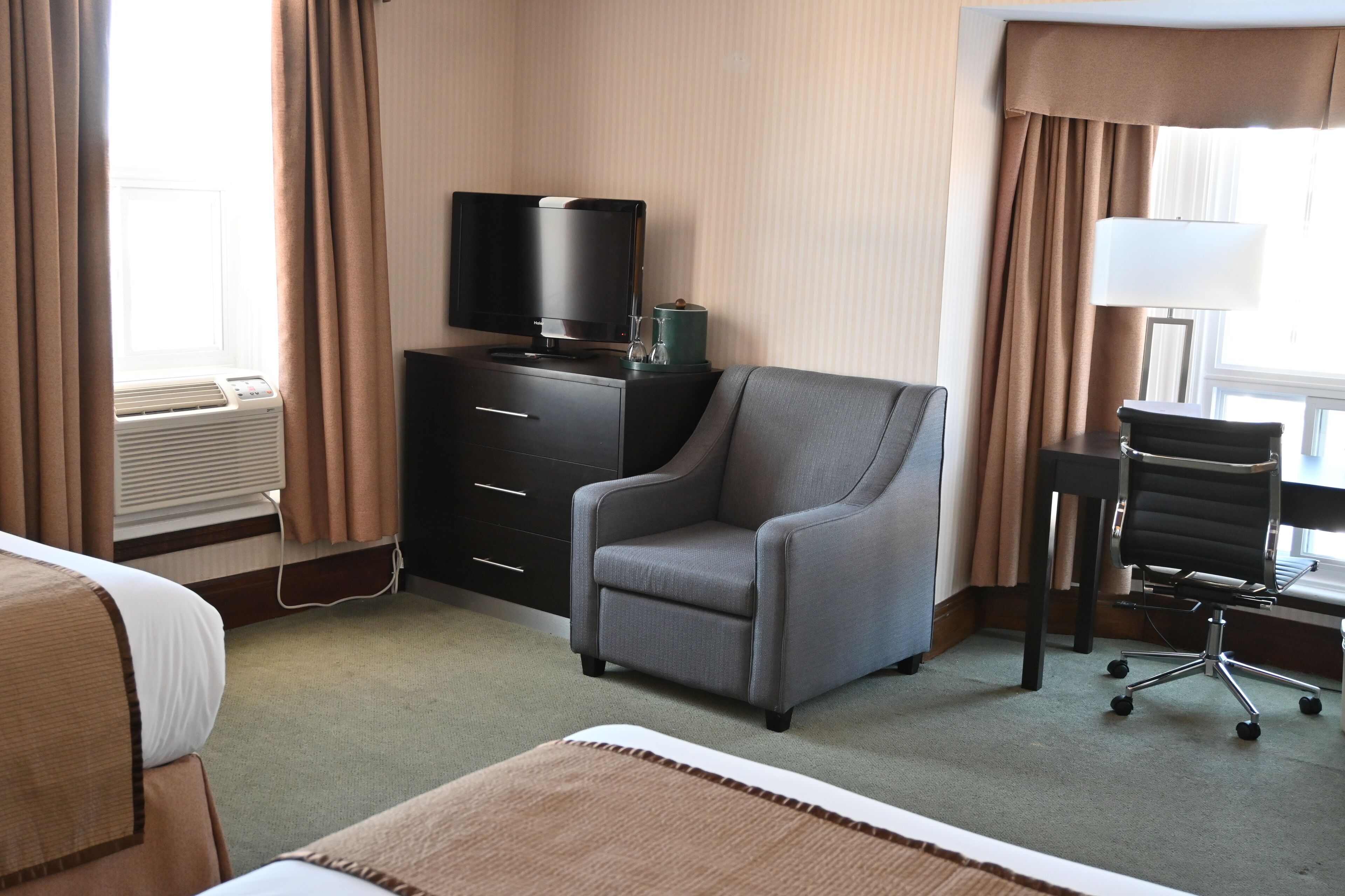 A hotel room with a bed, a desk with a computer, a chair, a TV on a dresser, and an air conditioning unit.