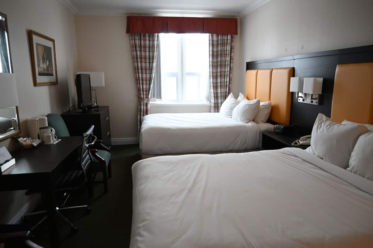 Quarto de hotel com duas camas, uma mesa de trabalho com cadeira, luminária e janela com cortinas.
