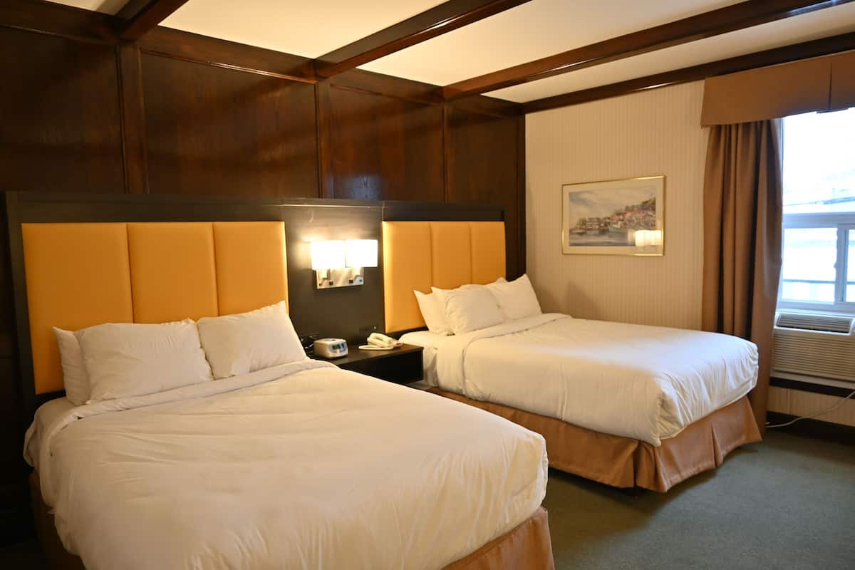 Quarto de hotel com duas camas, uma mesa de cabeceira, um abajur, um quadro e uma janela com cortinas.