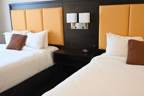 A hotel room with two beds, each with white linens and brown pillows, a nightstand with a clock, and wall-mounted lighting.