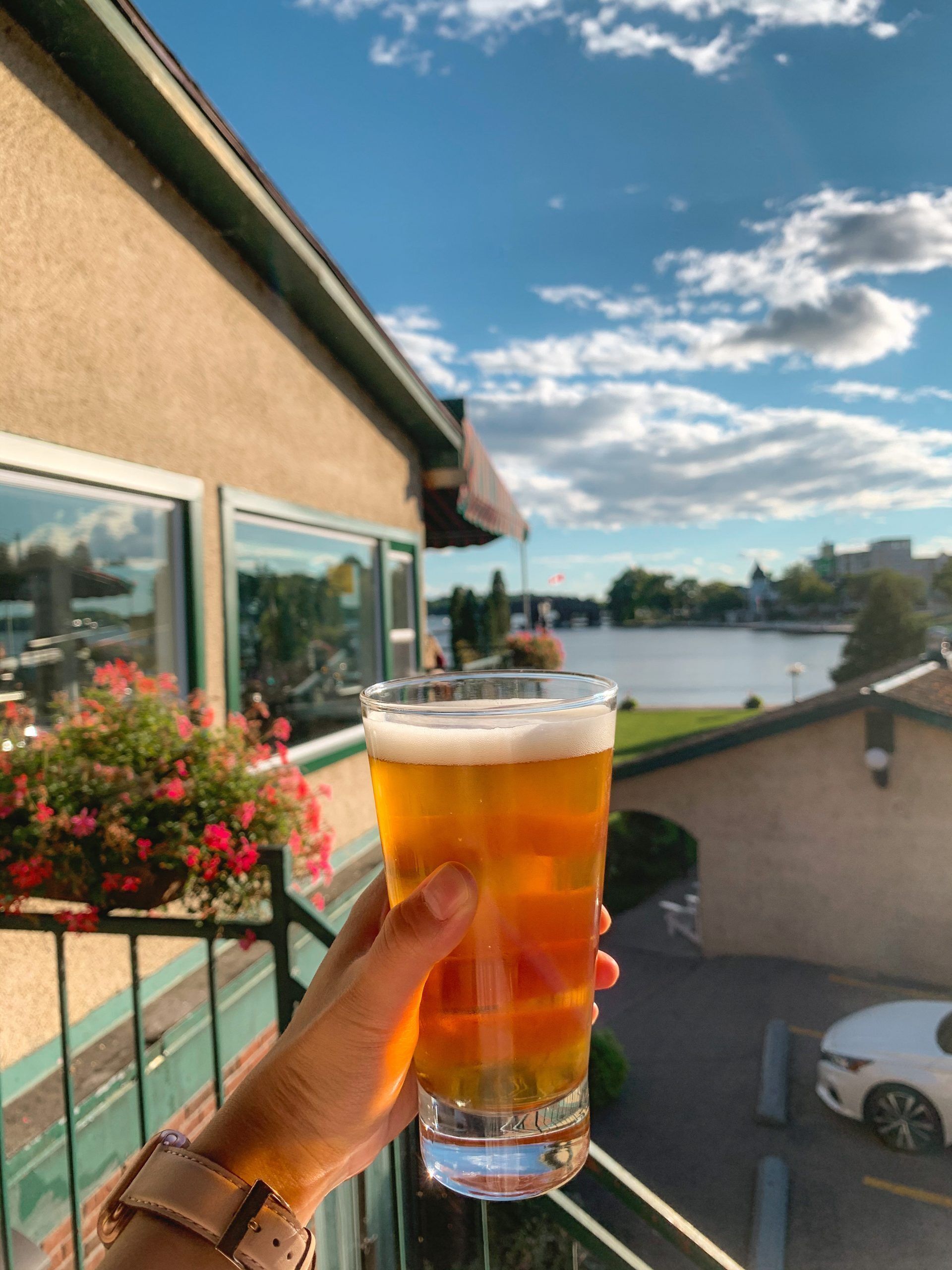 Una mà subjecta un got de cervesa, amb un edifici i un cos d'aigua al fons.