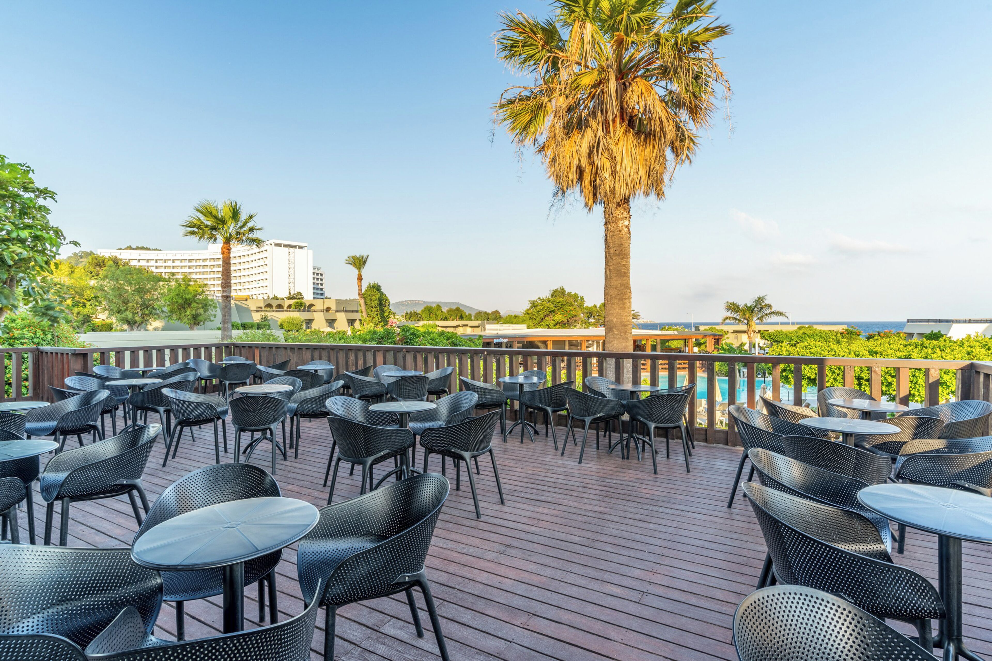 An outdoor patio with round tables and chairs, palm trees, and a view of a building and water in the background.