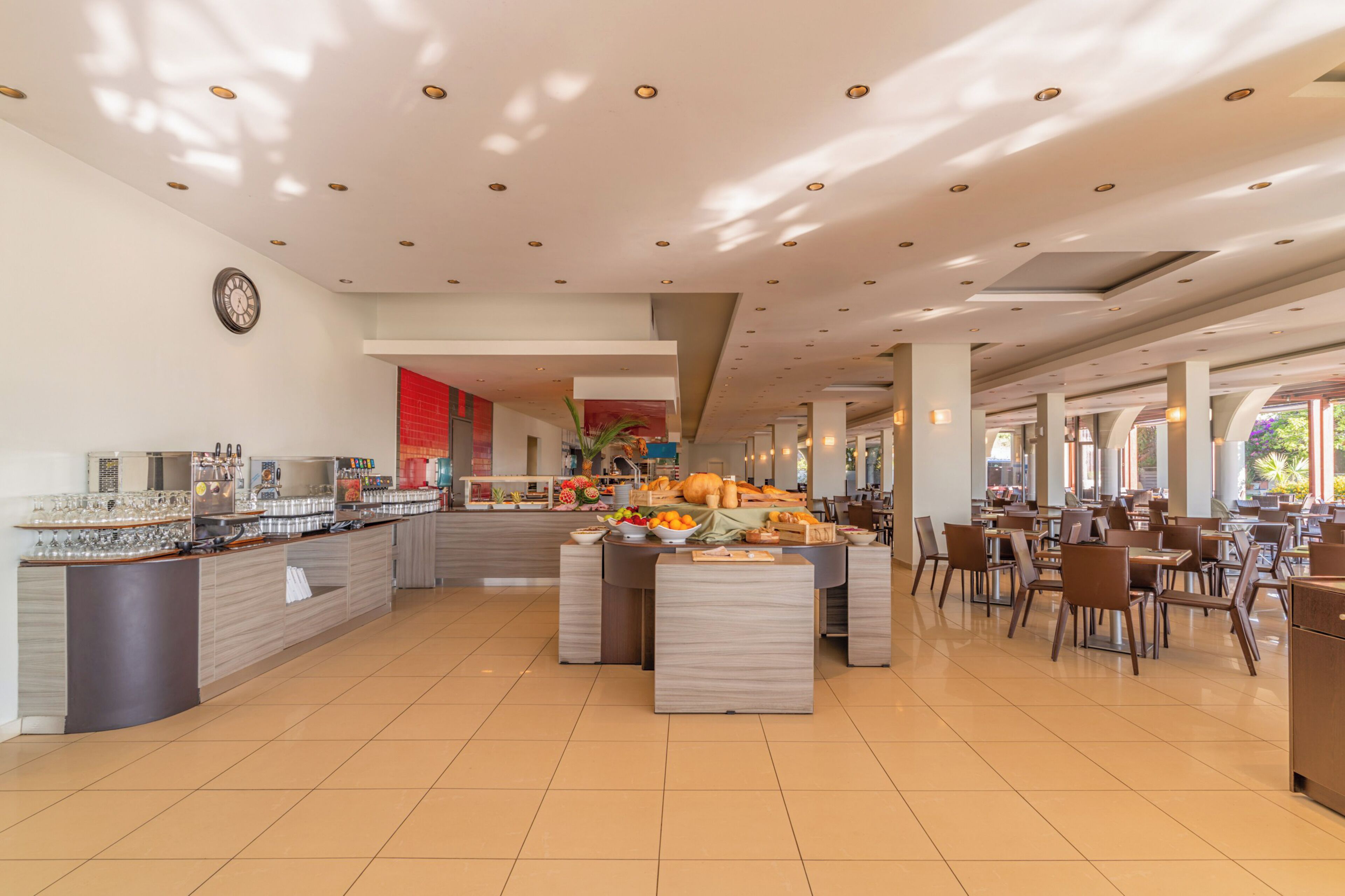 En rummelig hotelspisestue med en central buffet, forskellige siddepladser og et ur på væggen.