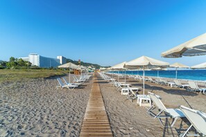 Una playa con filas de tumbonas y sombrillas, un paseo marítimo de madera y edificios al fondo.