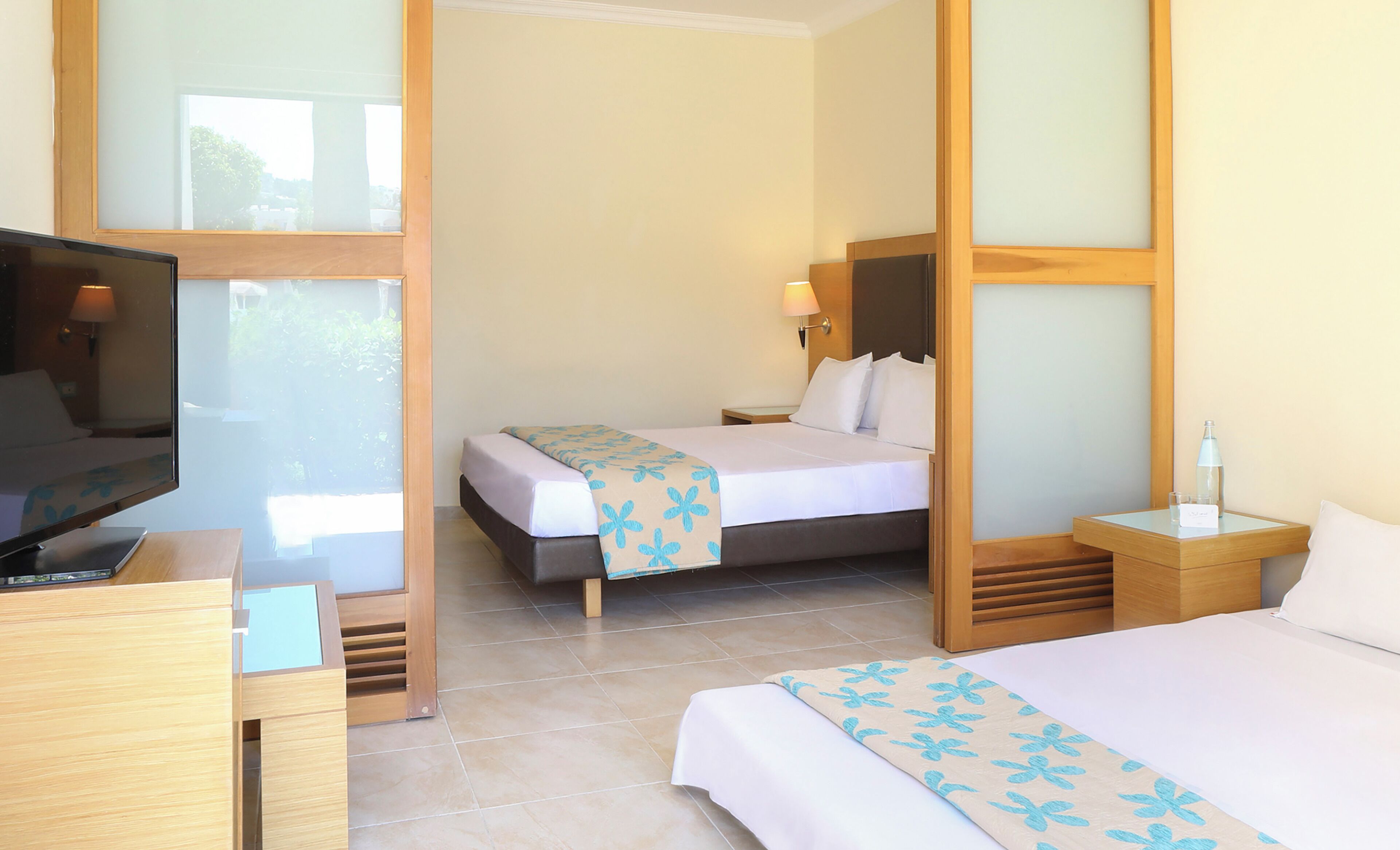 A hotel room with a bed, a desk with a TV, and a nightstand with a bottle and glasses.