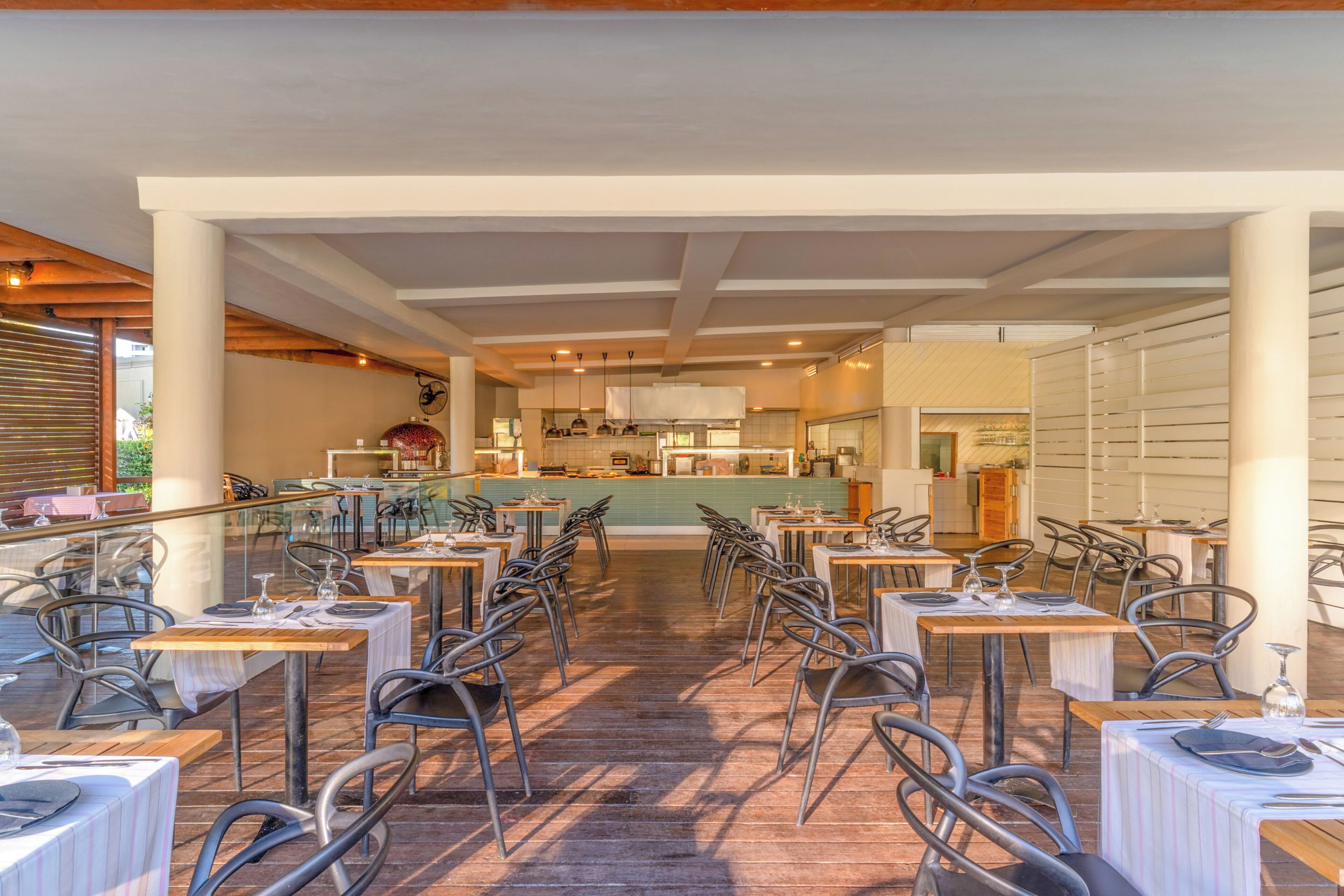 Un restaurante con varias mesas y sillas, una zona de cocina al fondo y vistas al exterior.