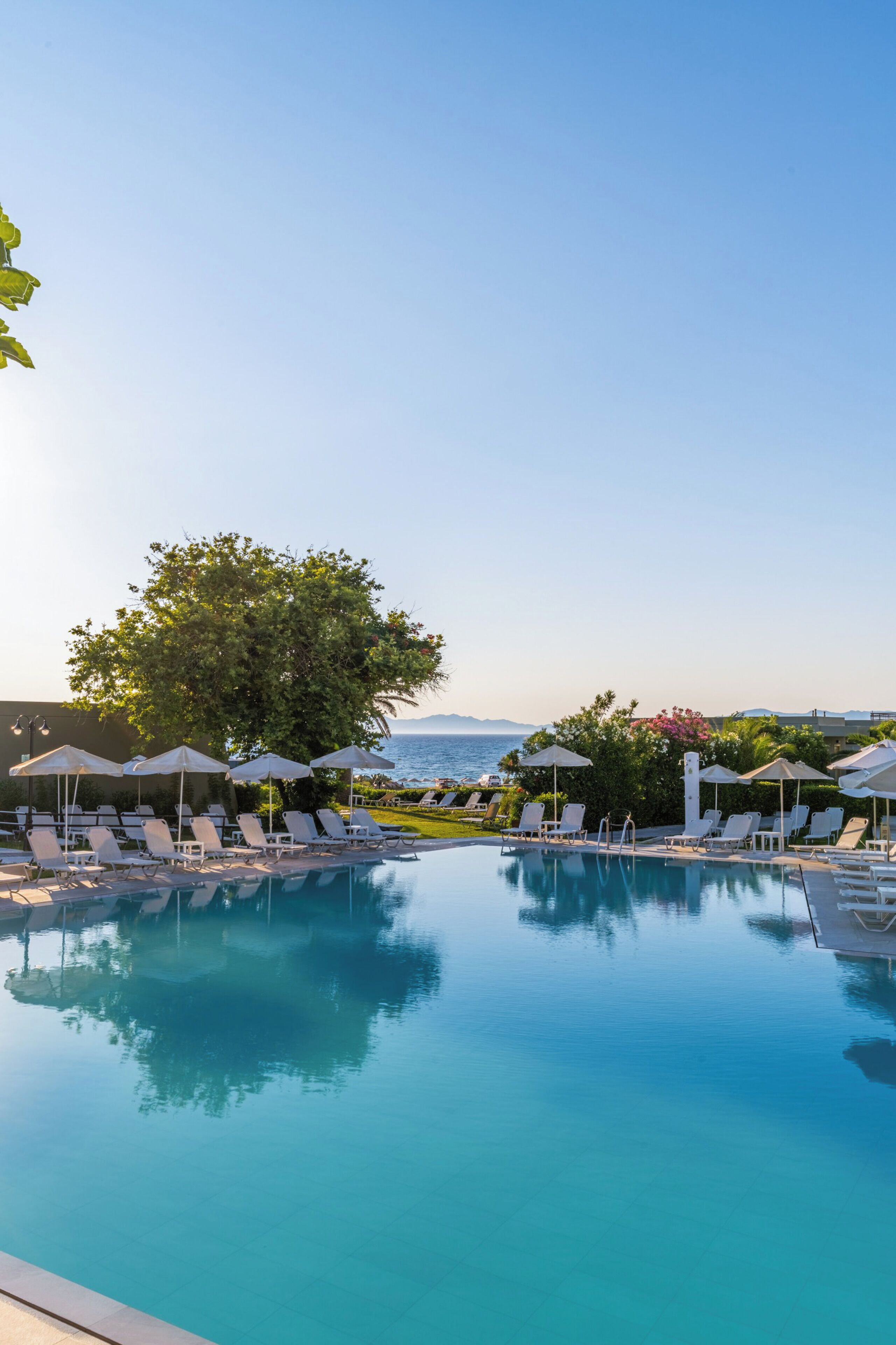 A pool with clear blue water, surrounded by white lounge chairs and umbrellas, with a view of the sea and mountains in the background.