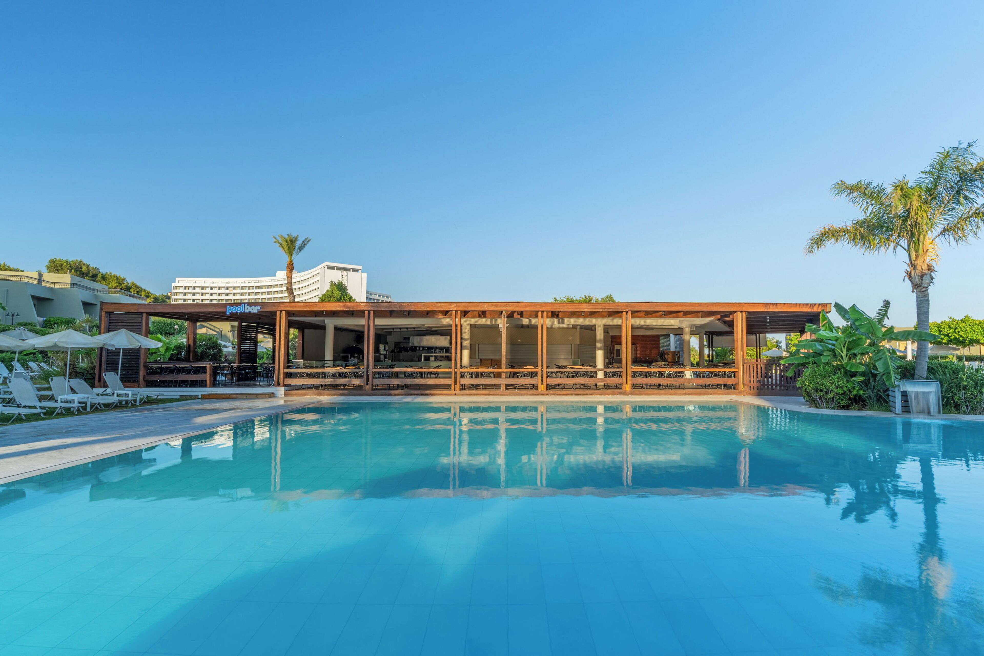 Un espace piscine comprenant un bar au bord de la piscine, des chaises longues et un bâtiment doté de grandes fenêtres.