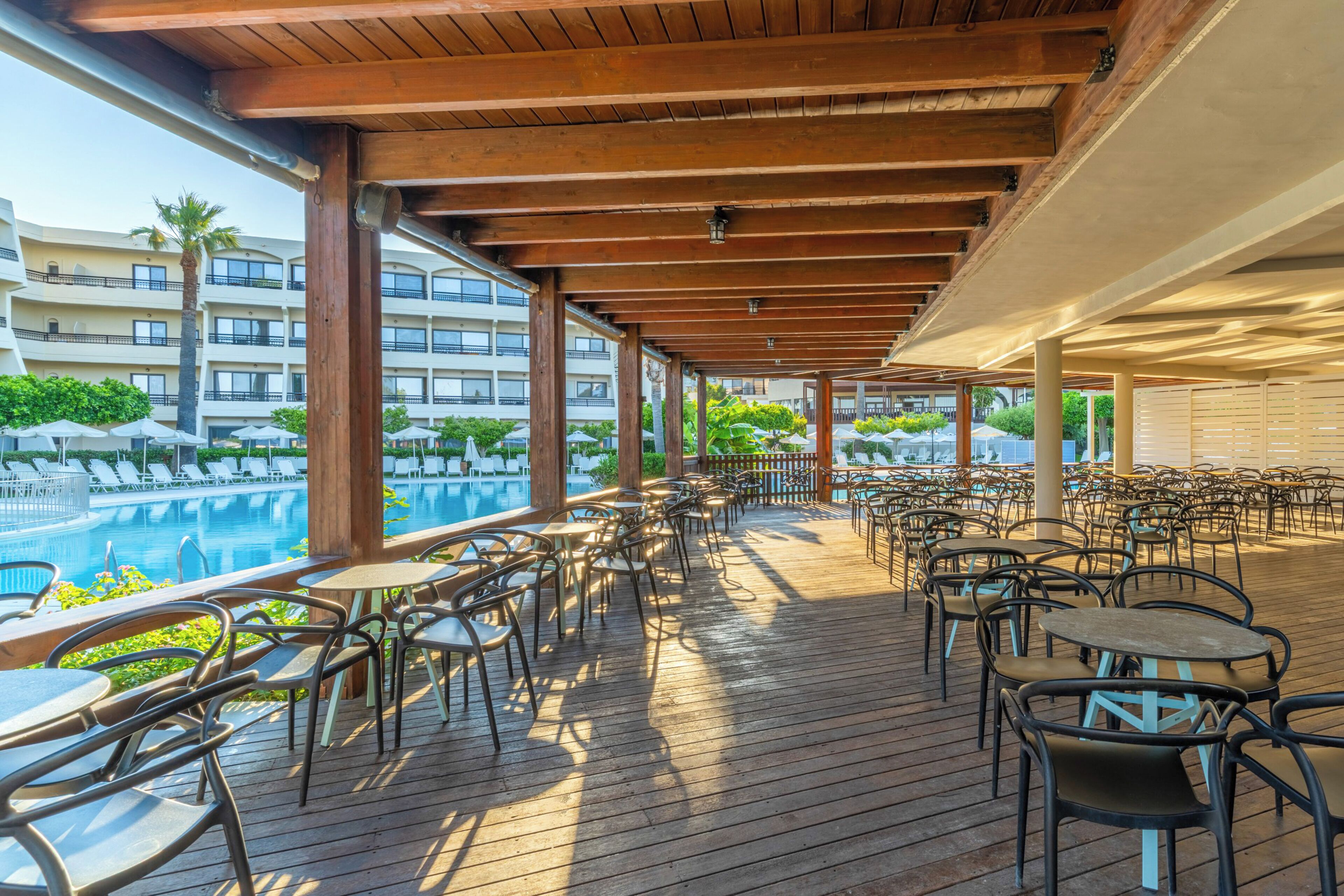 An outdoor deck with wooden flooring, metal chairs, and tables, overlooking a swimming pool and a hotel building.