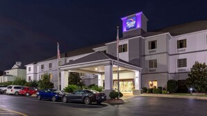 A Sleep Inn hotel with a covered entrance, a parking lot with several cars, and a flagpole with an American flag. - Sleep Inn & Suites Millbrook - Prattville (Millbrook)