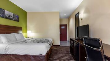 A hotel room with a bed, a desk with a chair, a television, and a door in the background.