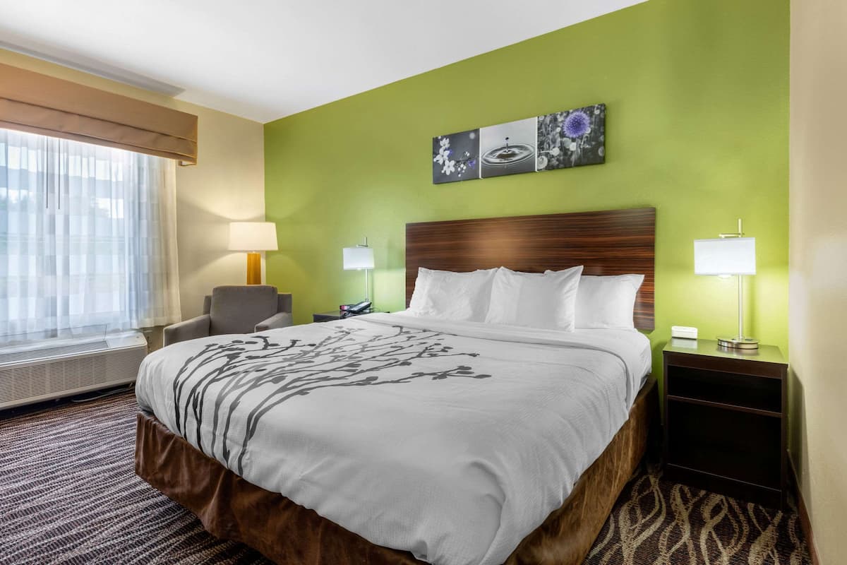 A hotel room with a large bed, two bedside tables with lamps, a chair, a window with curtains, and a wall with three framed pictures.
