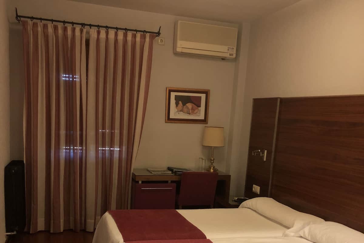 A hotel room with a bed, a desk, a chair, a lamp, an air conditioner, and a curtain.