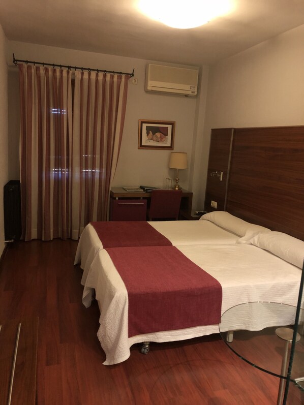A hotel room with a bed, a desk, a chair, a lamp, an air conditioner, and a curtain.
