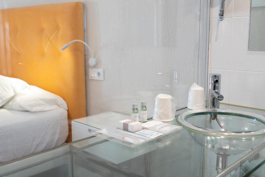Habitación de hotel con mesita de noche de cristal, cama con ropa de cama blanca, lavamanos con estructura de cristal y un espejo.