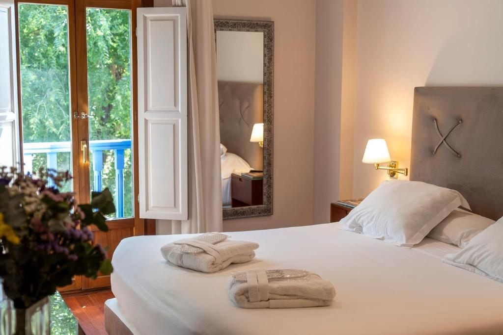 A hotel room with a bed, two bedside lamps, a mirror, and a window with a view of greenery.