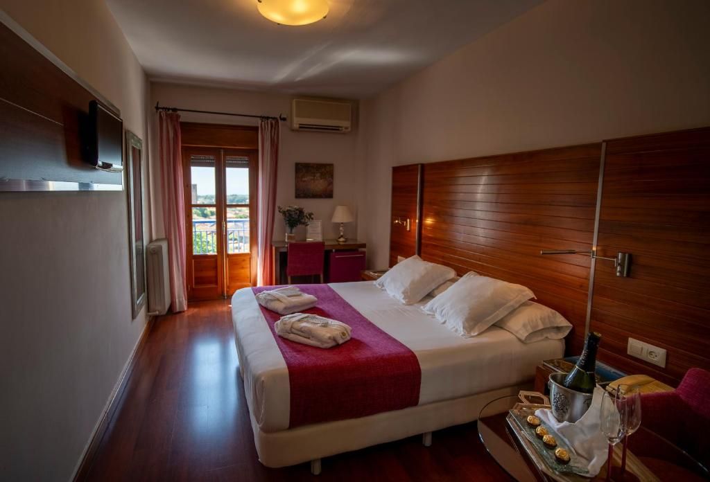 Ein Hotelzimmer mit einem großen Bett, einem Holzkopfende, Blick auf einen Balkon, einer Minibar und einem Fernseher.