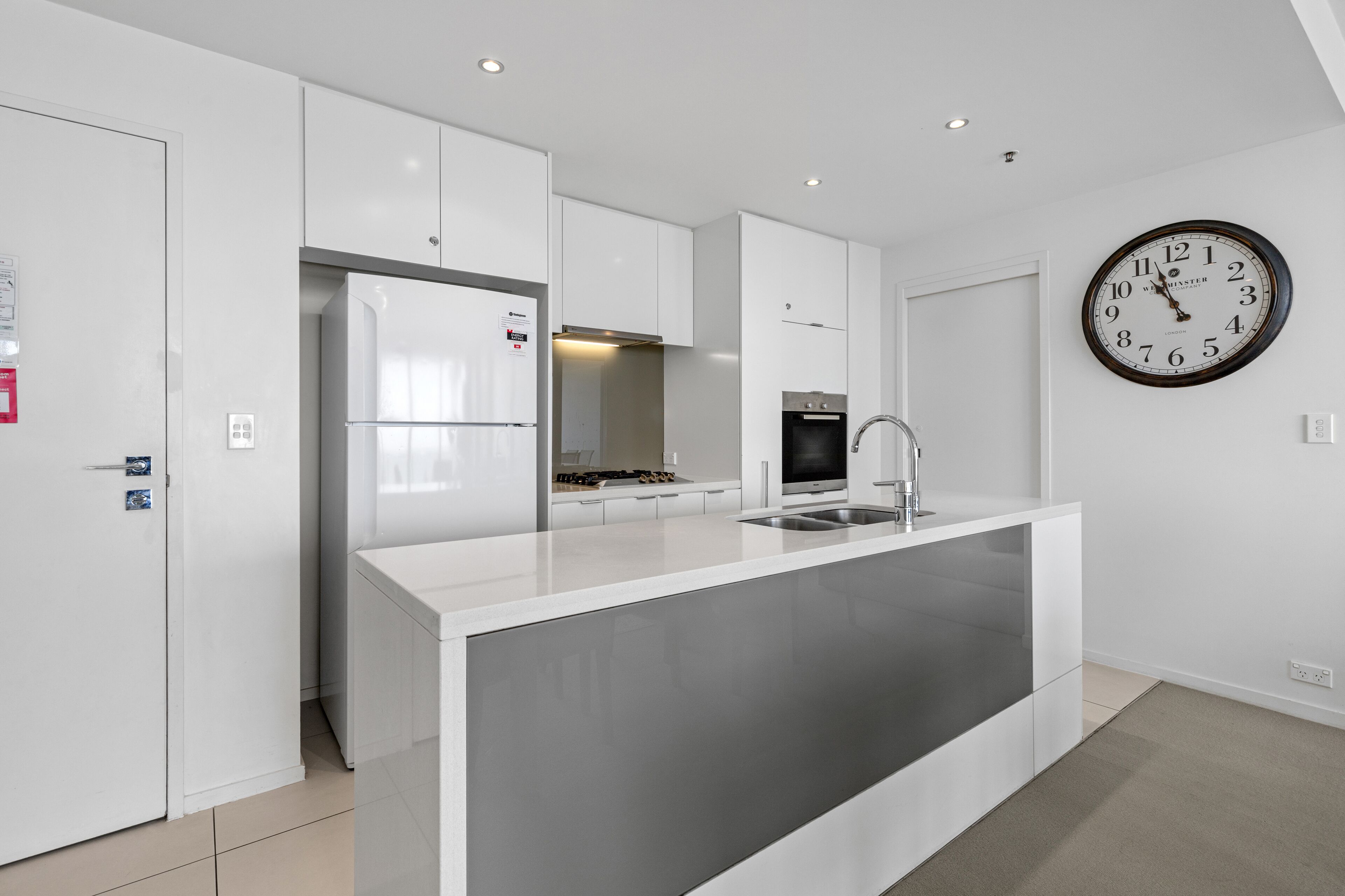 A modern kitchen with white cabinetry, a central island with a sink, a built-in oven, and a wall-mounted clock.