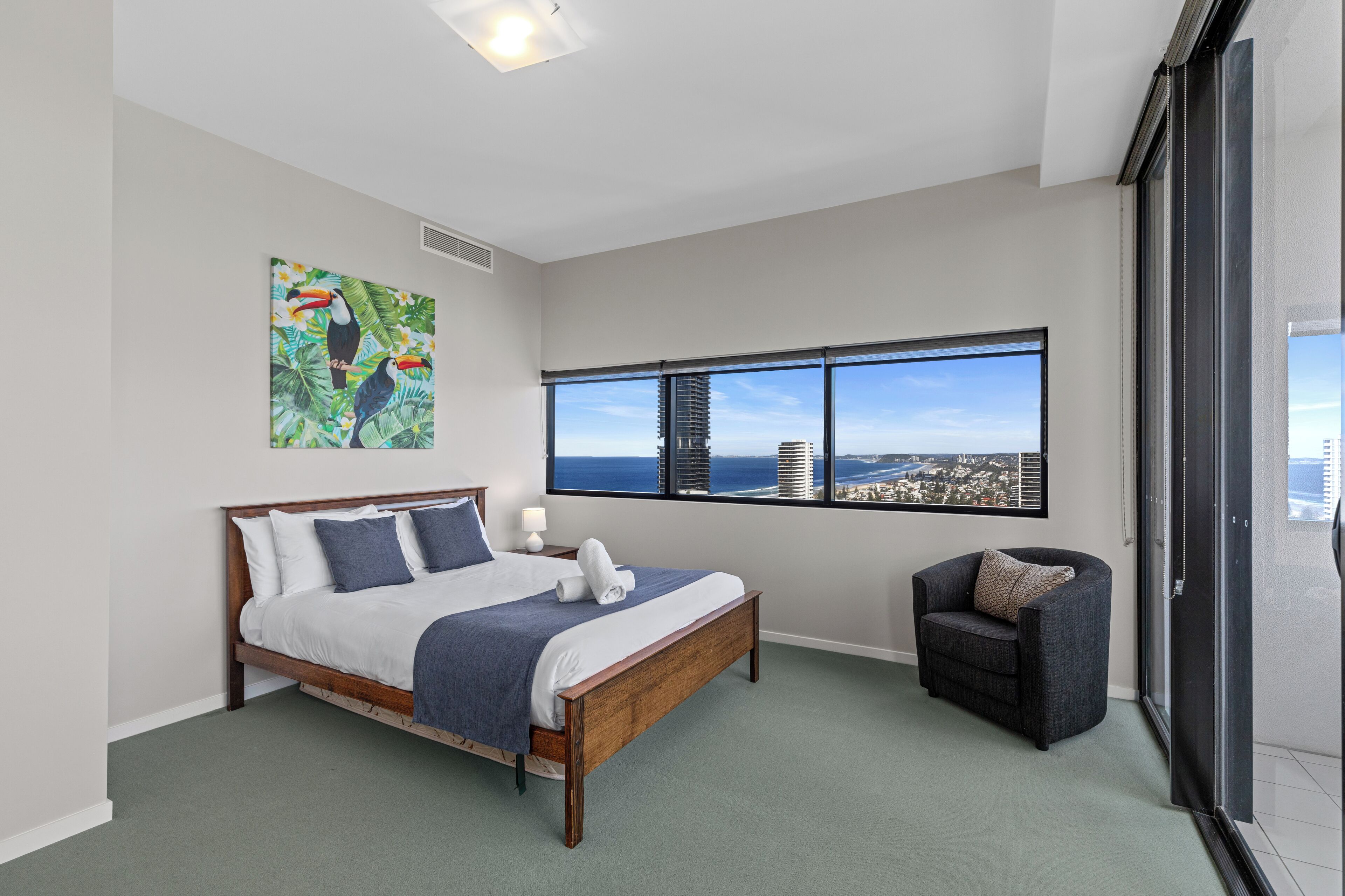 A bedroom with a large bed, a chair, a window with a city view, and a tropical-themed painting.