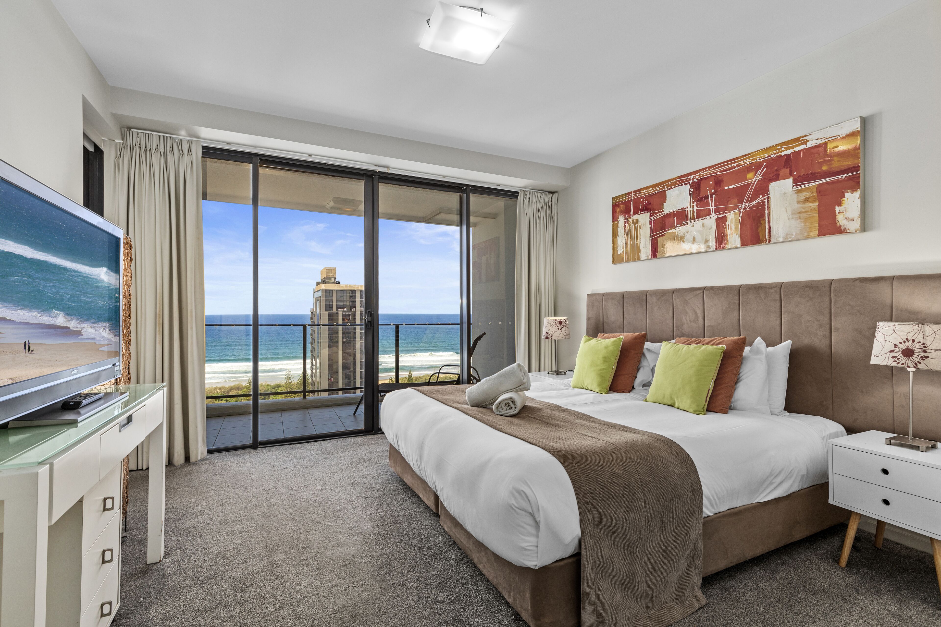A hotel room with a large bed, a TV, a desk, and a balcony with a view of the ocean.