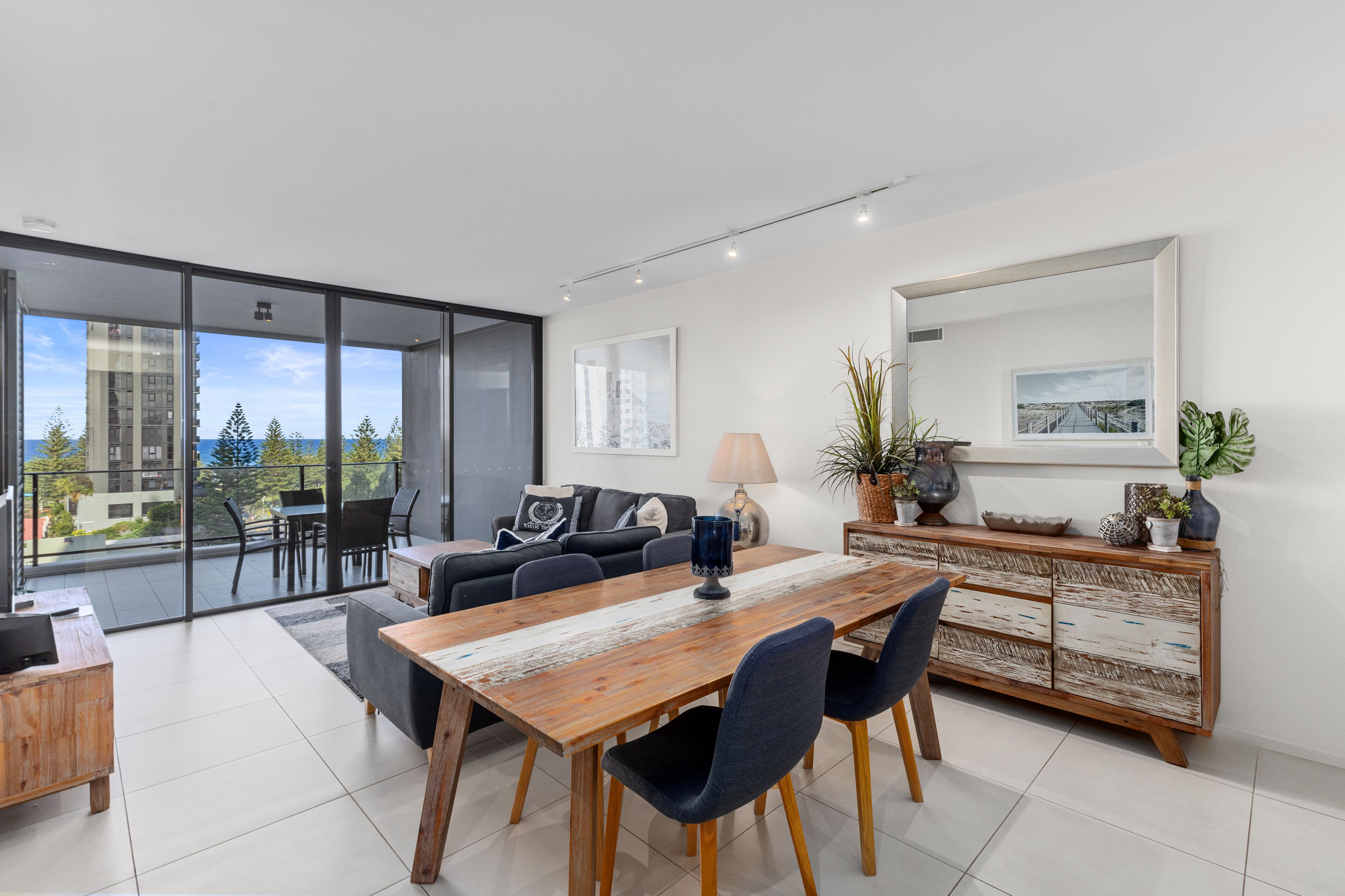 A modern living room with a wooden dining table, blue chairs, a sofa, and a large window overlooking a cityscape.