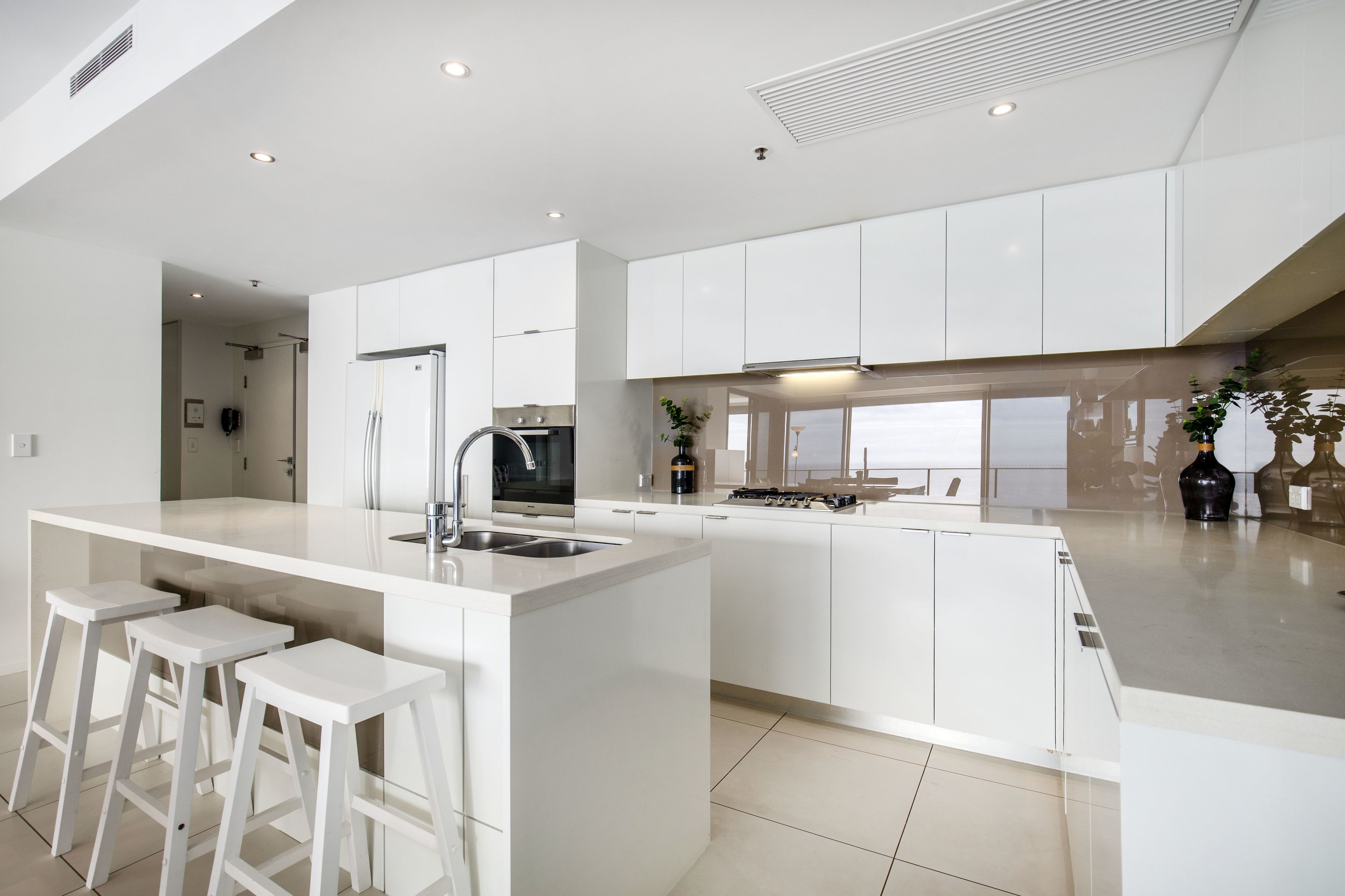 A modern kitchen with white cabinetry, a central island with stools, a built-in oven, and a large window providing natural light.