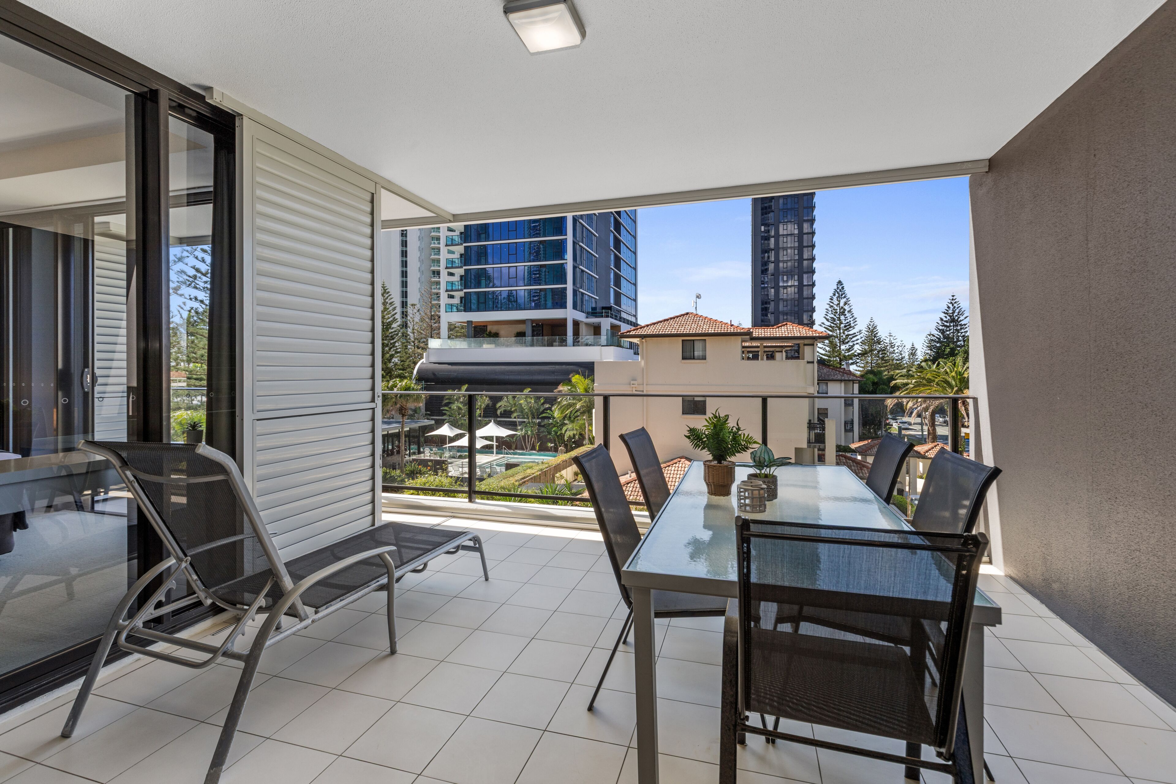 A balcony with a glass table and chairs, overlooking a pool and buildings.
