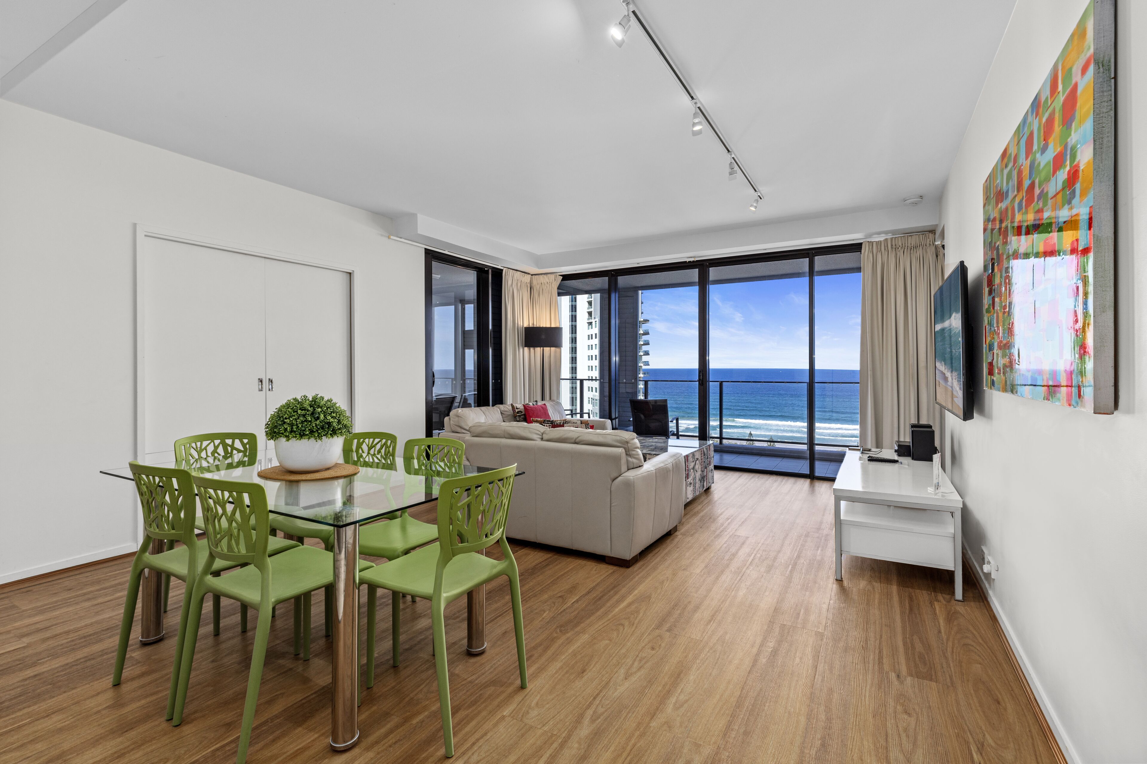 A modern living room with a glass dining table, green chairs, a beige sofa, and a large window offering a view of the ocean.