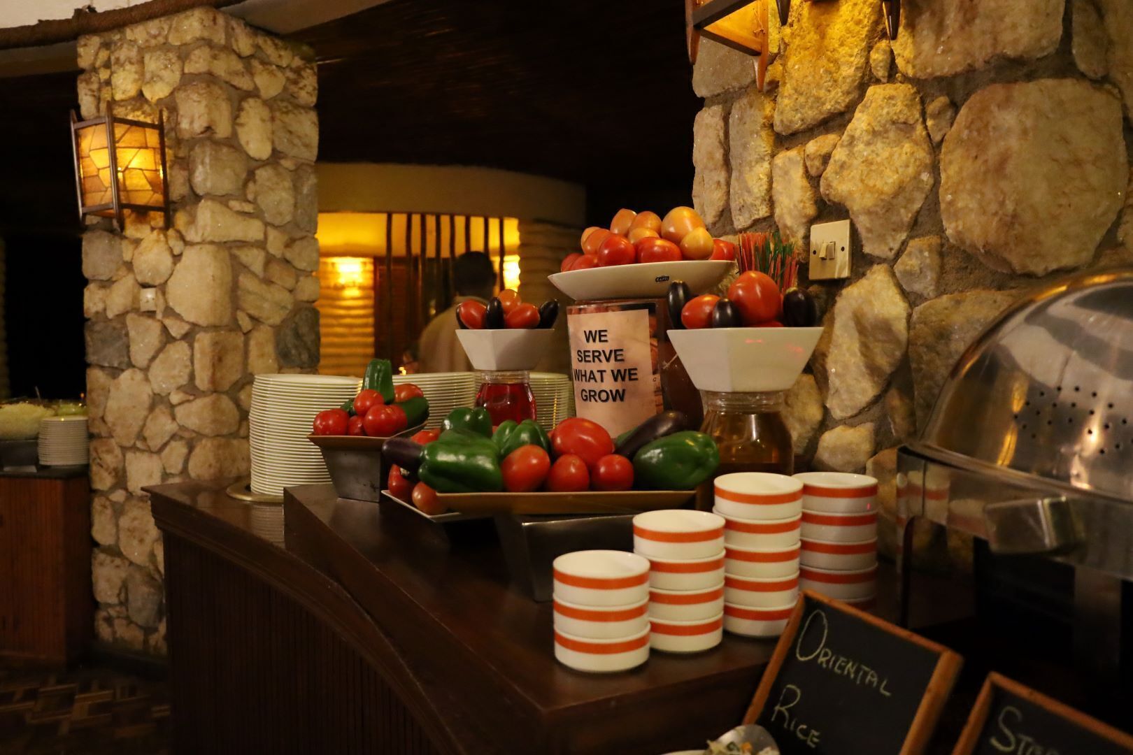 A buffet area with a stone wall backdrop, a variety of fresh vegetables, and labeled serving dishes.