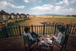 Dos personas sentadas en sillas junto a una mesa con bebidas y un libro, con vistas a un campo que incluye animales y edificios al fondo.