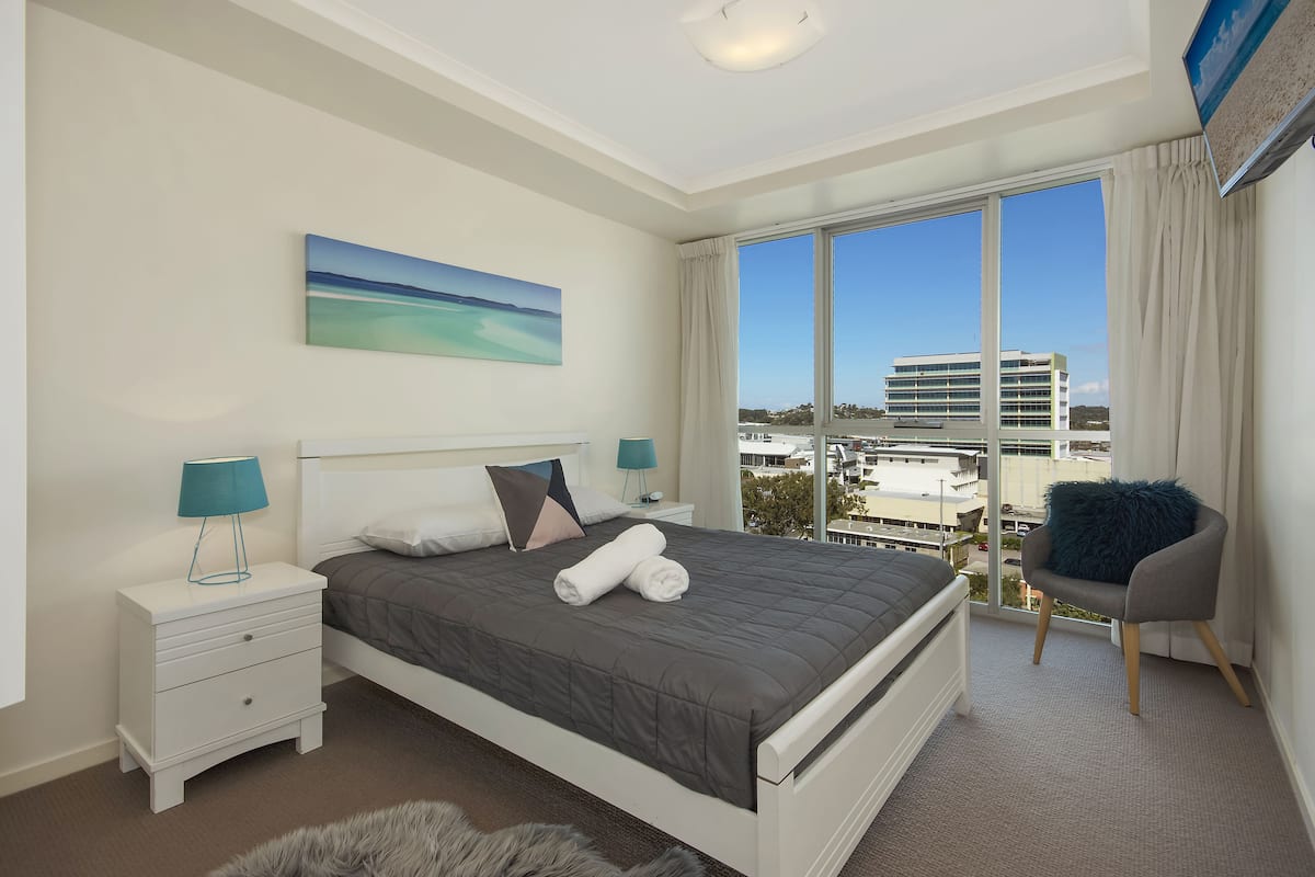 A modern bedroom with a large bed, a nightstand, a chair, and a view of the city.