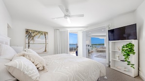 Un dormitorio con una cama grande, un ventilador de techo, un televisor sobre un soporte blanco y vista al océano a través de una puerta abierta.