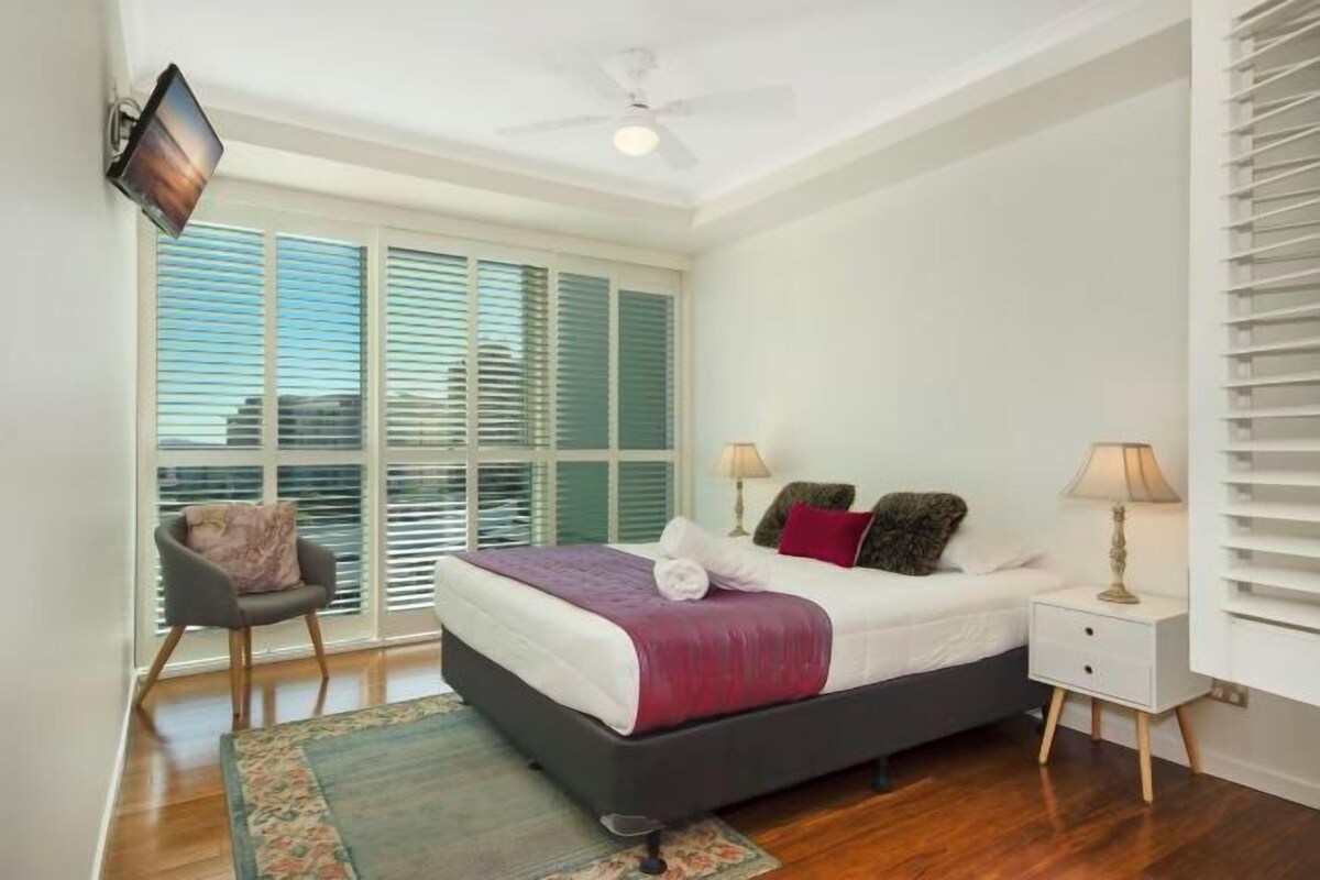 A bedroom with a bed, bedside tables, a chair, a ceiling fan, and a window with blinds.