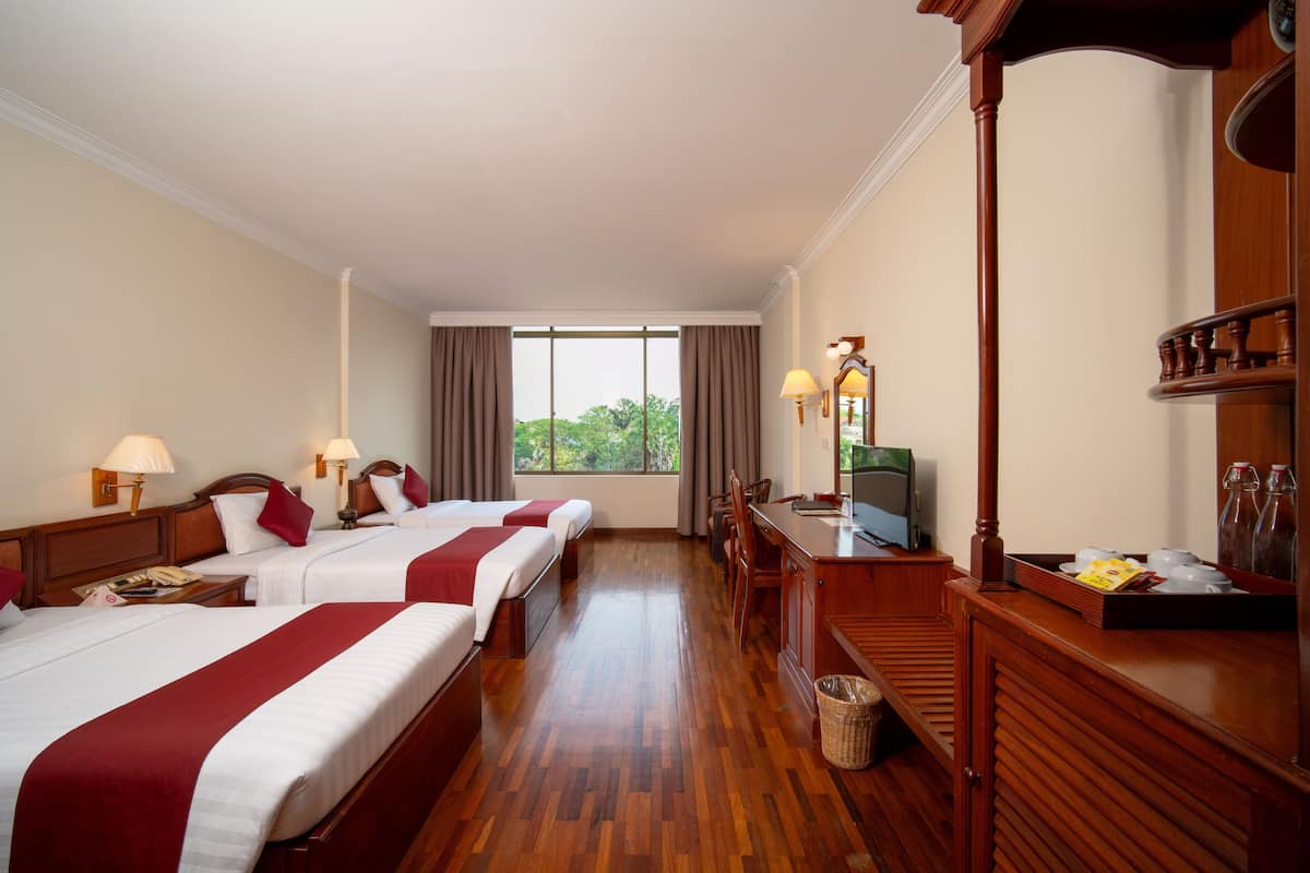 A hotel room with two beds, a desk with a TV, a wooden dresser, and a window with a view of trees.