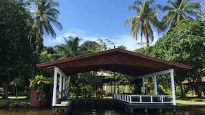 A covered pavilion with a wooden roof and white columns, situated over a body of water, surrounded by lush greenery and palm trees.