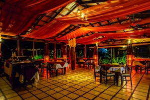 Un comedor interior con mesas y sillas, piso de baldosas y techo con telas colgantes.