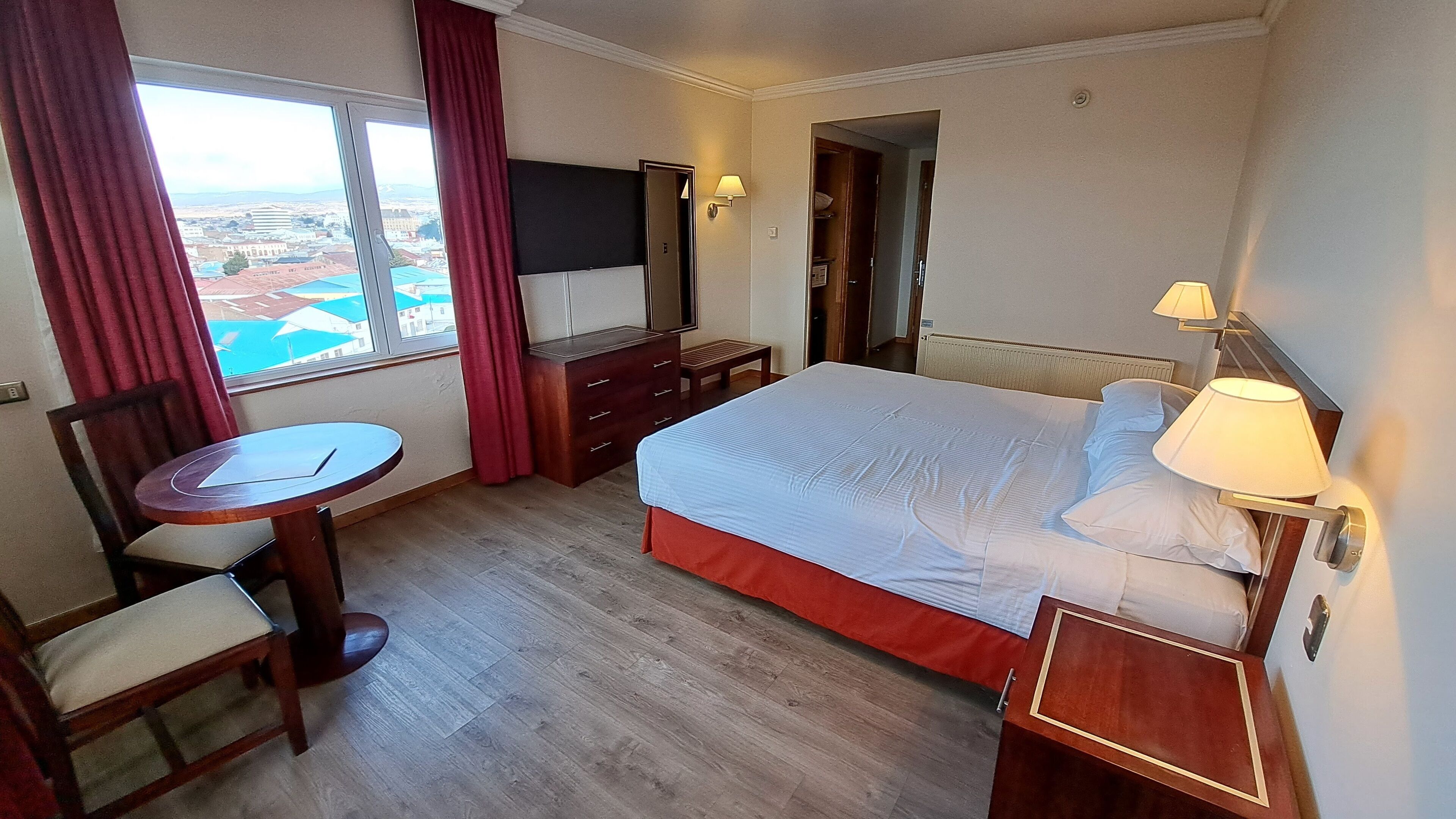Una habitación de hotel con cama, escritorio, silla, televisión y ventana con cortinas.